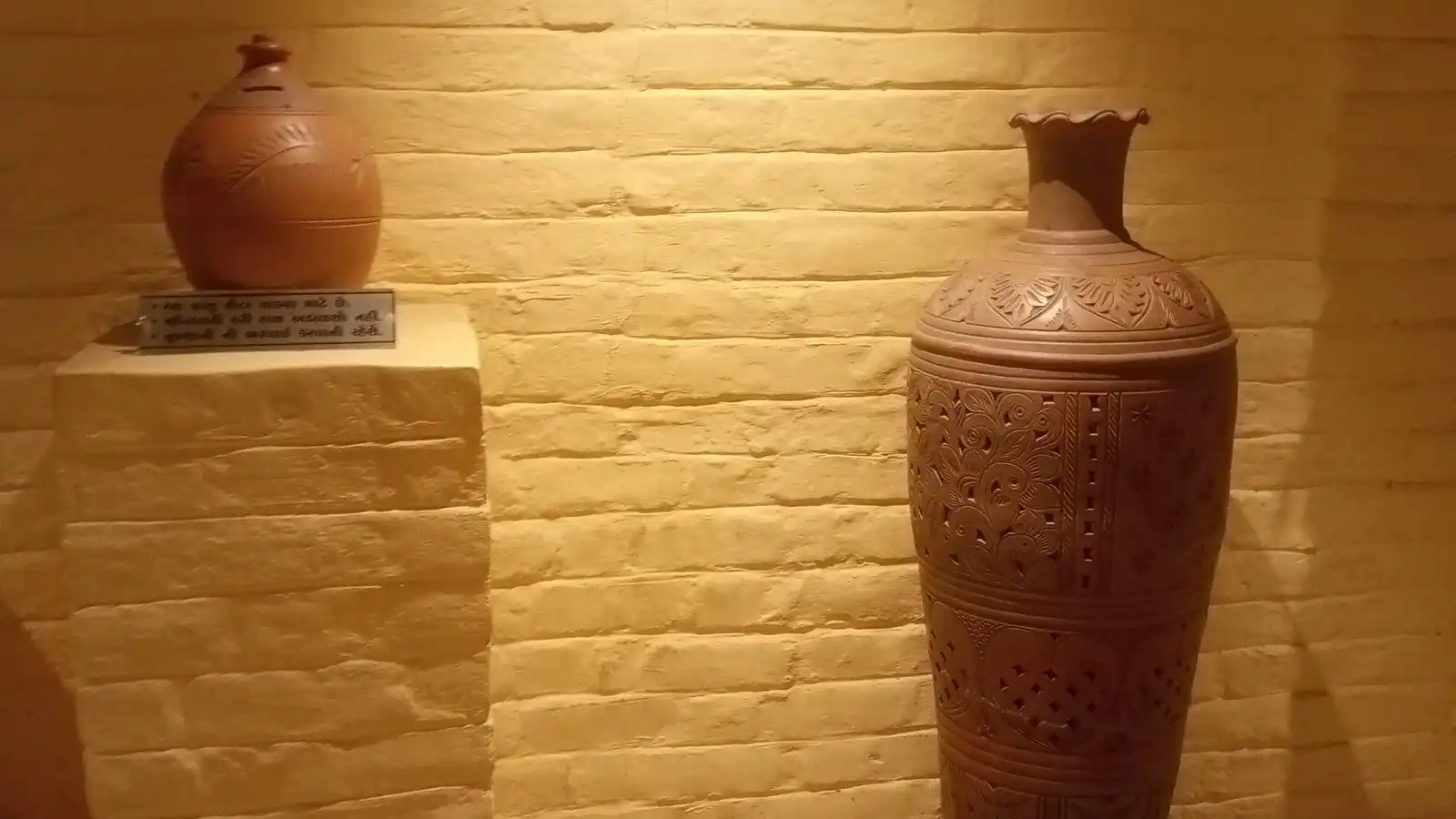 Two clay vases displayed on a plinth and the floor against a lit yellow brick wall