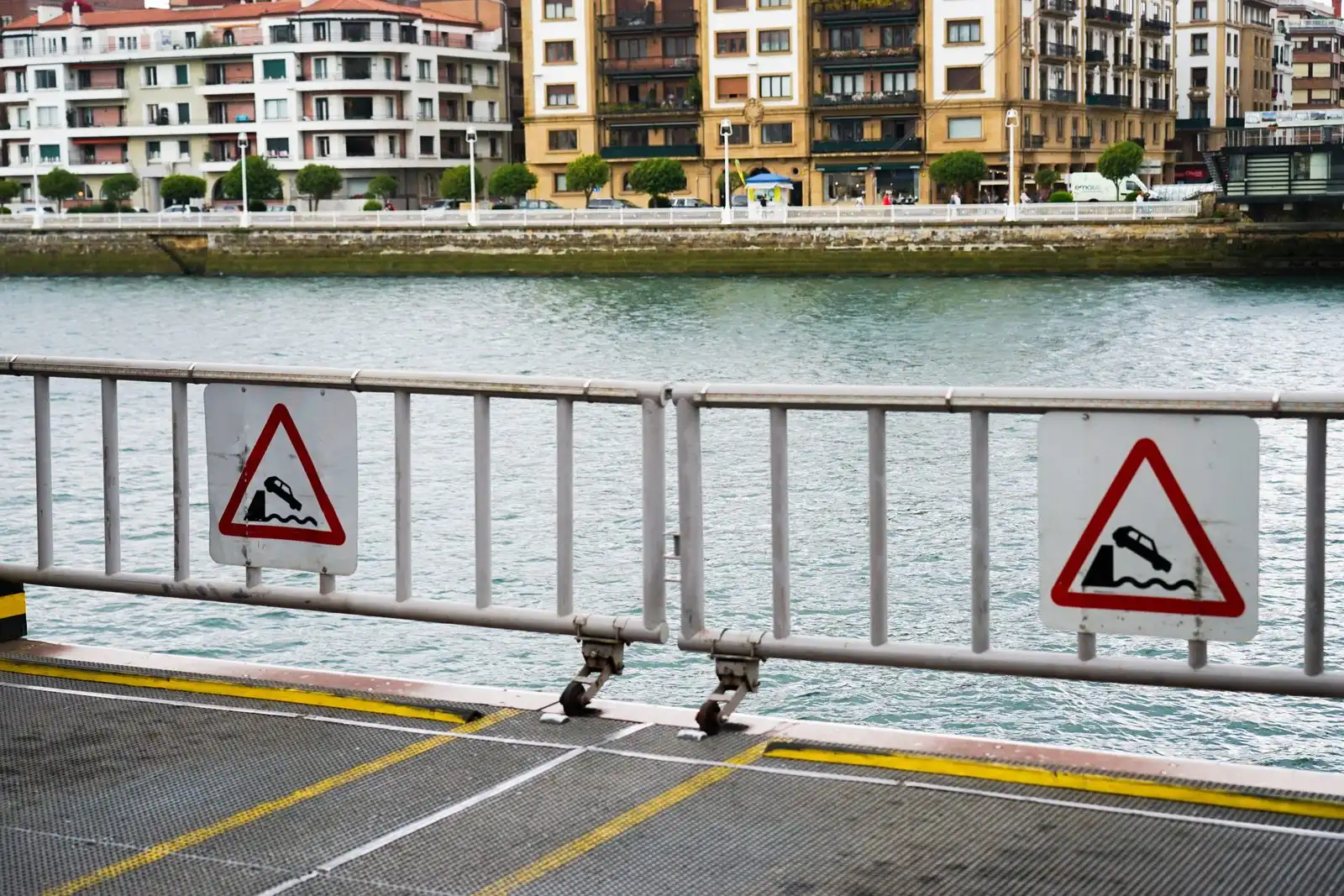 A ferry ramp barrier with two red-bordered warning signs, a river and apartment blocks behind