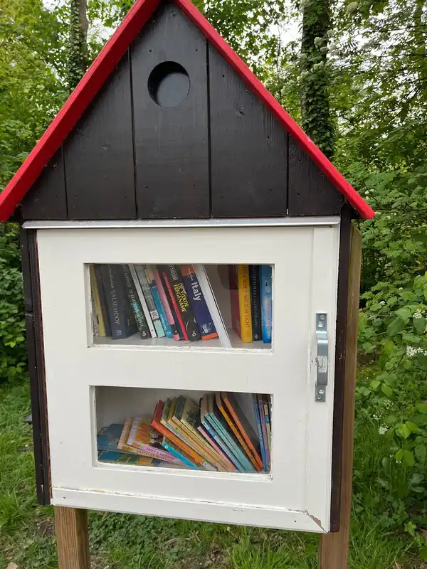 A small wooden book-exchange box with a red roof, full of books, on a post in a garden