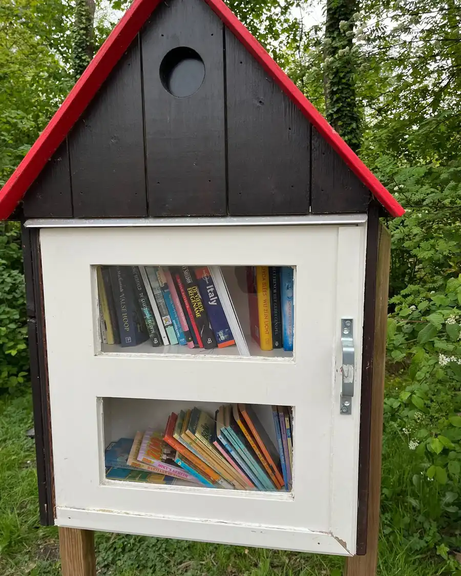 A small wooden book-exchange box with a red roof, full of books, on a post in a garden