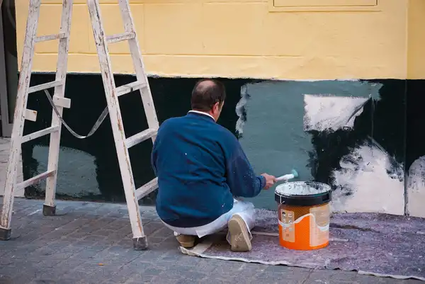 A painter kneeling on a sheet, painting a dark green panel on a wall below a ladder