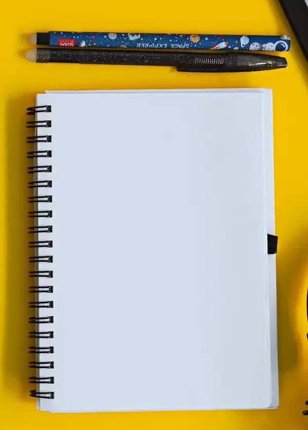 A blank spiral notebook and two pens on a bright yellow background